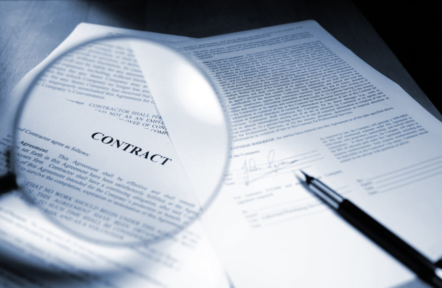 A close up blue toned image of a magnifying glass held out in front of page in a legal contract document. Sheets of paperwork are on a wooden desk with a ink pen close to a signed page. Contract text and signature are fake. Shot with  shallow depth of field with focus on the word ‘Contract’.